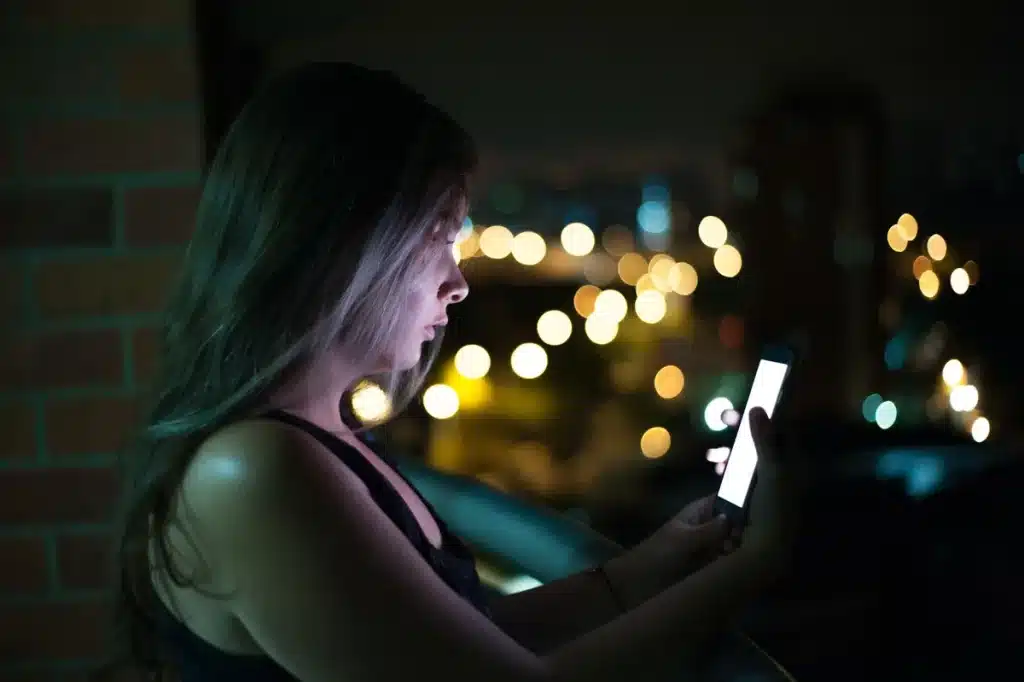 Person sitzt nachts allein vor einem leuchtenden Smartphone-Bildschirm in dunklem Zimmer.